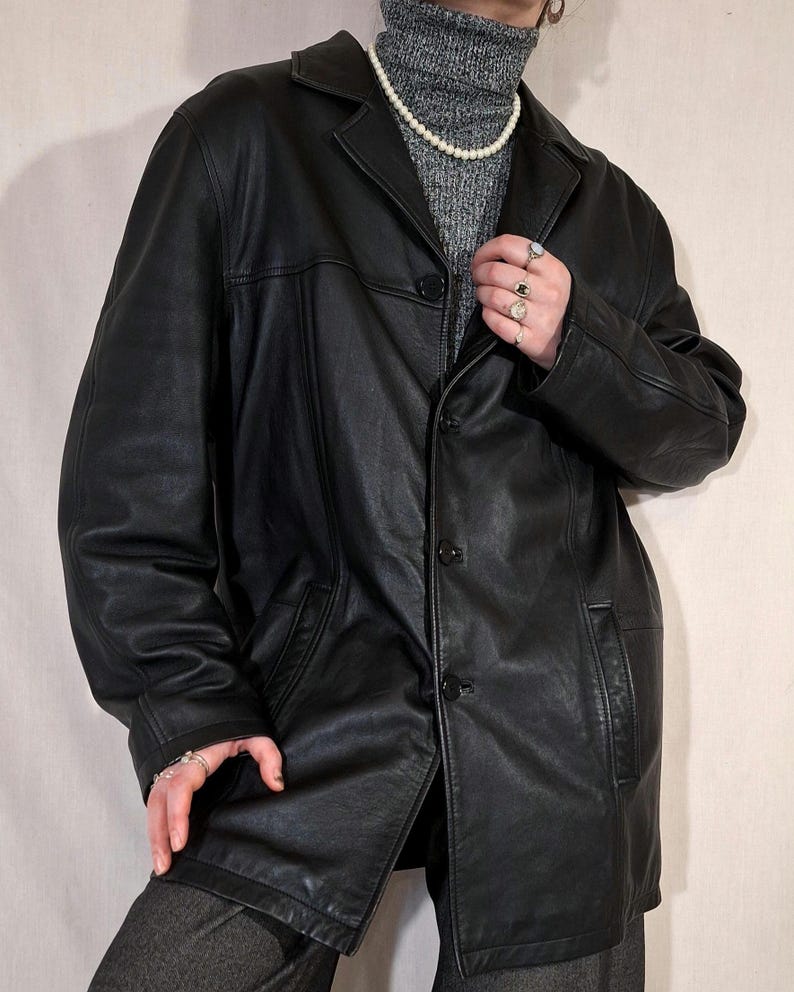 May include: A black leather jacket with a classic design. The jacket features a collared neckline, button-down closure, and front pockets. The jacket is worn over a gray turtleneck sweater and gray pants.