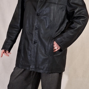 May include: A black leather jacket with a button-up front and a collared neckline. The jacket has two front pockets and is worn over a gray turtleneck sweater. The person is wearing gray pants.