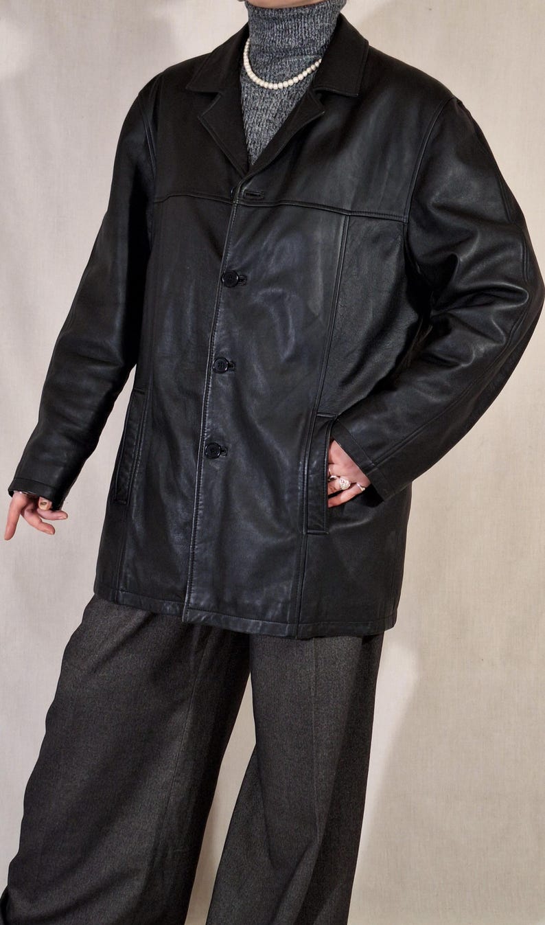 May include: A black leather jacket with a button-up front and a collared neckline. The jacket has two front pockets and is worn over a gray turtleneck sweater. The person is wearing gray pants.