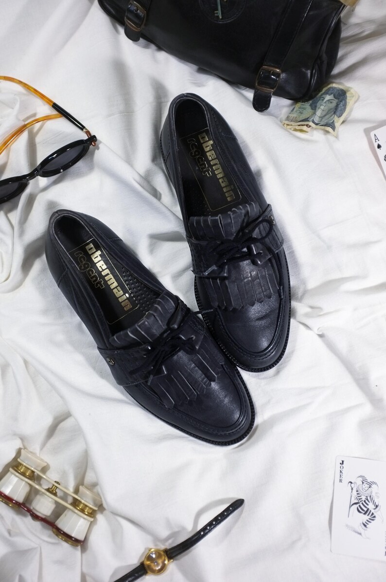 May include: A pair of black leather tassel loafers with a gold label that reads "Obermain Regent".