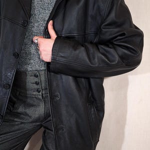 May include: A black leather jacket with a classic lapel collar and button closure. The jacket is worn over a gray turtleneck sweater and gray trousers. A pearl necklace is visible.