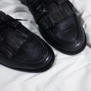 May include: A pair of black leather loafers with fringe detailing and black laces. The loafers are on a white surface.