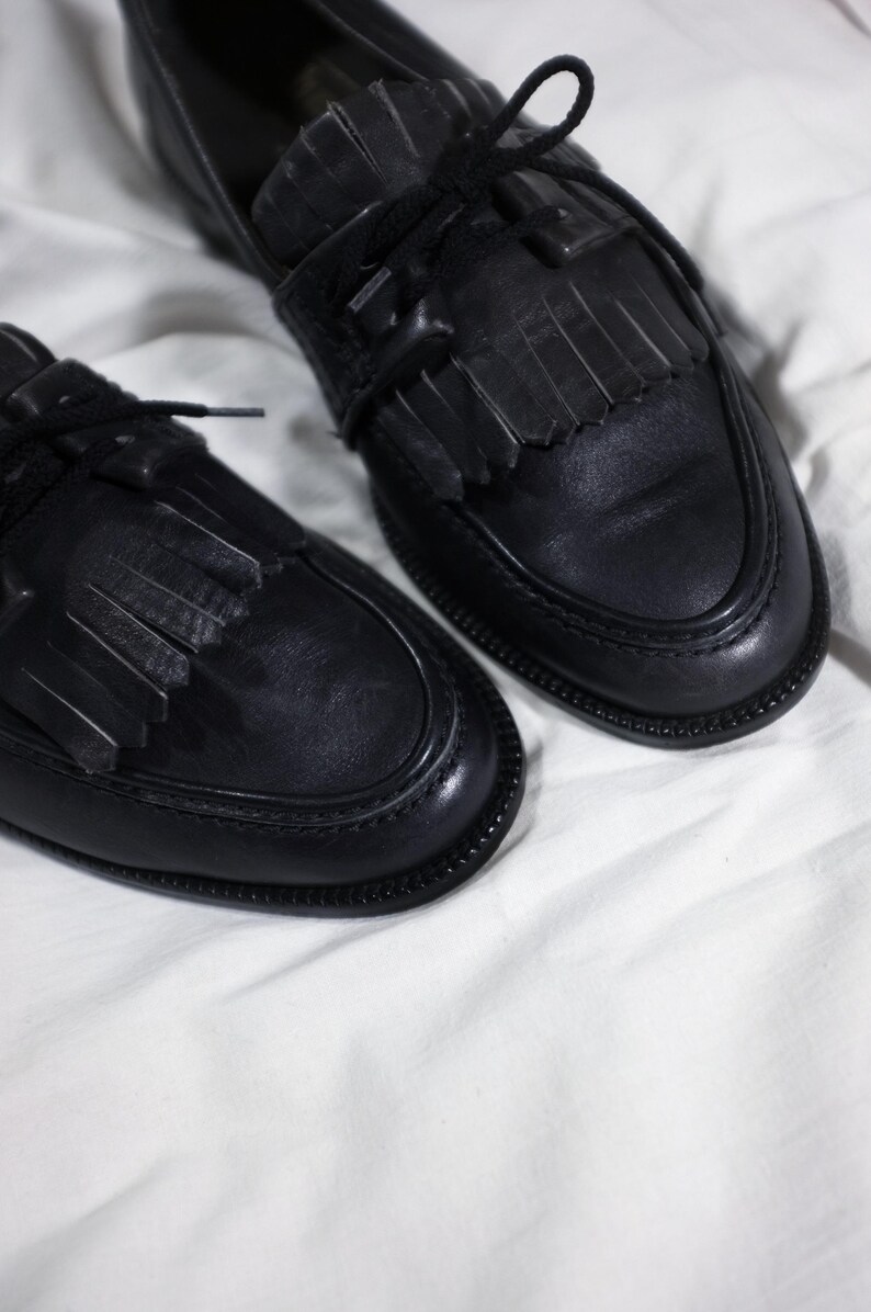 May include: A pair of black leather loafers with fringe detailing and black laces. The loafers are on a white surface.