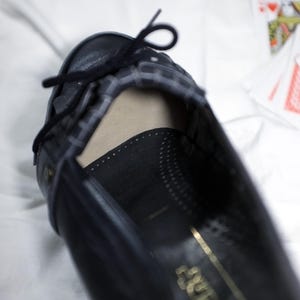 May include: A black leather shoe with a gold inscription that reads "Aldo" on the sole. The shoe has a black ribbon tied around the ankle.