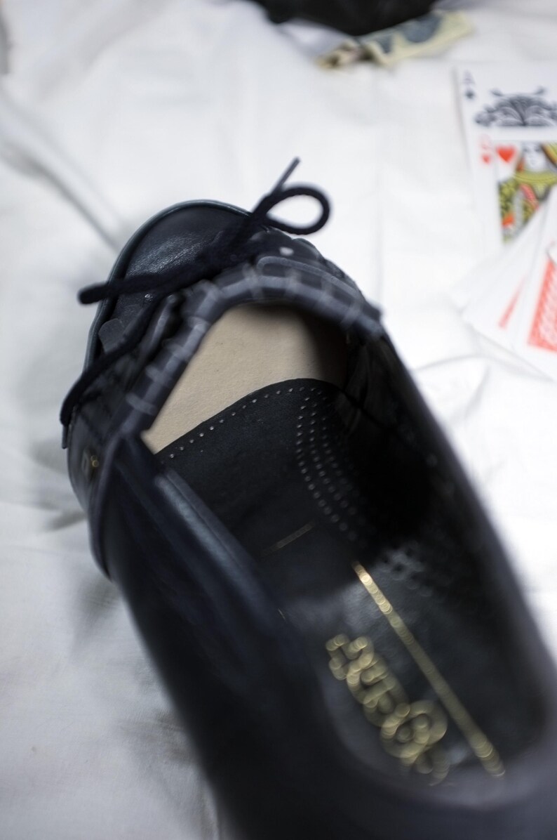 May include: A black leather shoe with a gold inscription that reads "Aldo" on the sole. The shoe has a black ribbon tied around the ankle.