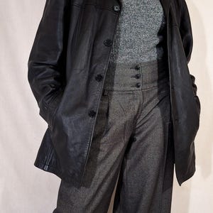 May include: A black leather blazer with a classic collar and button closure. The blazer is worn over a gray turtleneck sweater and high-waisted gray trousers. A pearl necklace is visible.