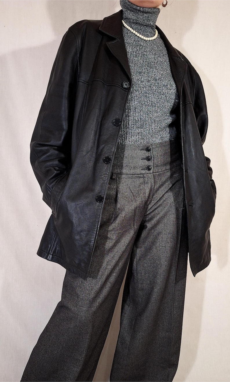 May include: A black leather blazer with a classic collar and button closure. The blazer is worn over a gray turtleneck sweater and high-waisted gray trousers. A pearl necklace is visible.