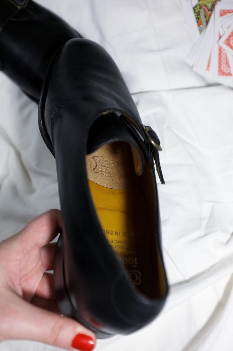 May include: Black leather dress shoe with a buckle closure. The shoe has a yellow leather insole with the text "Goodrich" and "Established Since 1902" printed on it. The text "Made in England" is also printed on the insole.
