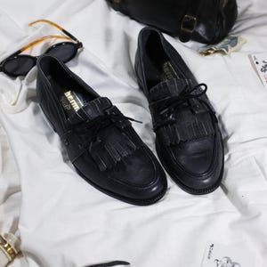 May include: A pair of black leather tassel loafers with a black lace-up closure. The shoes are on a white surface.