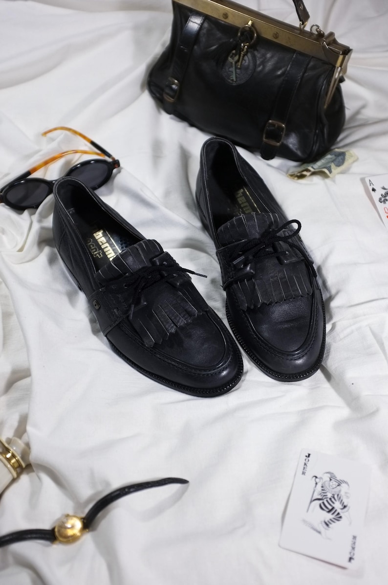 May include: A pair of black leather tassel loafers with a black lace-up closure. The shoes are on a white surface.