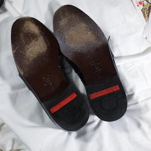 May include: A pair of black leather shoes with red rubber soles. The shoes have a worn look and are showing signs of wear.