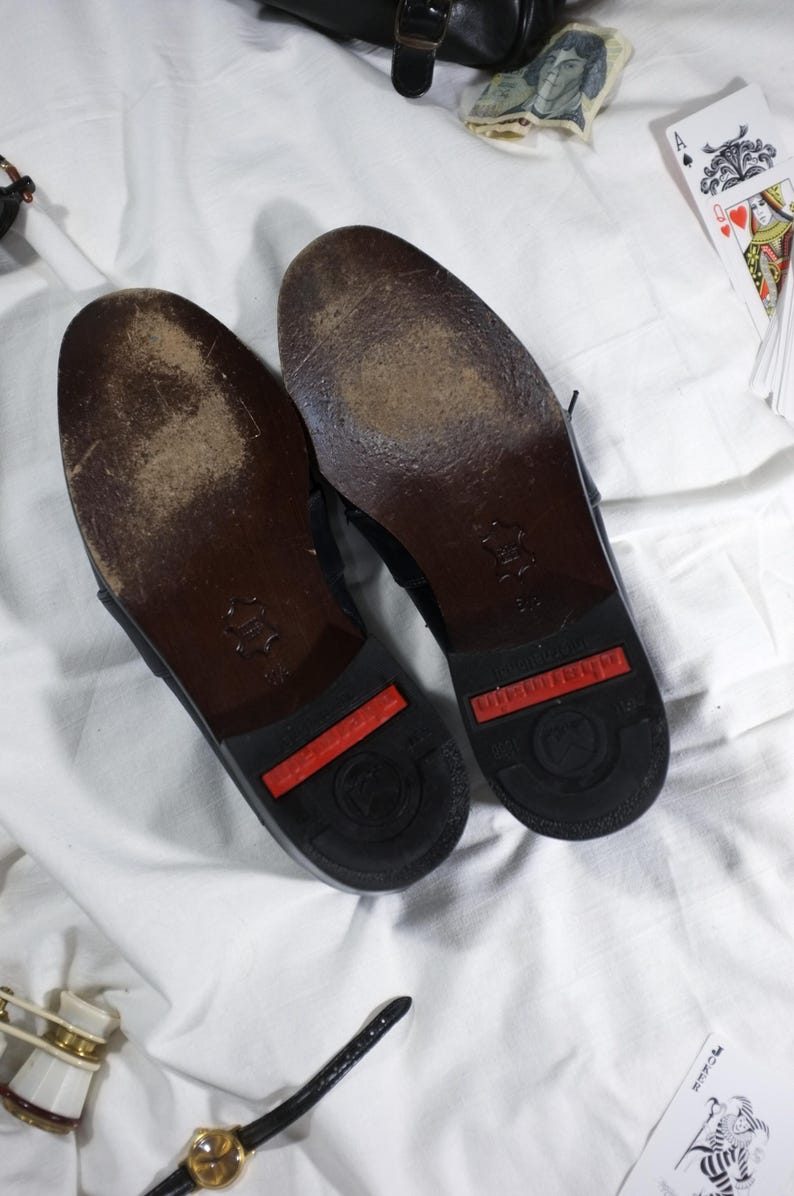 May include: A pair of black leather shoes with red rubber soles. The shoes have a worn look and are showing signs of wear.