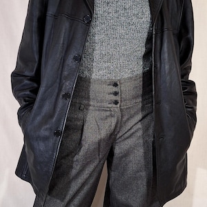 May include: A black leather blazer over a gray turtleneck sweater and high-waisted gray trousers. A pearl necklace is visible. The outfit is a mix of textures and tones, creating a classic, layered look.