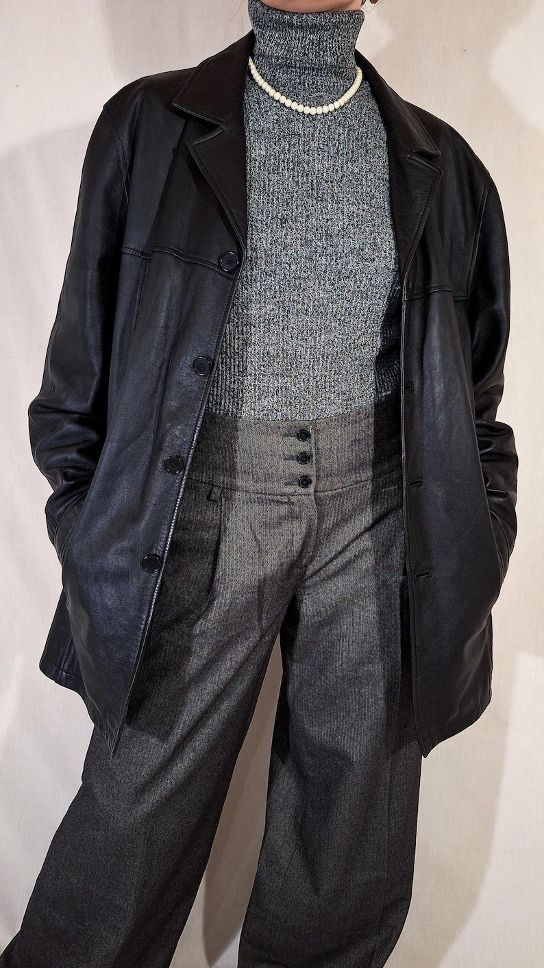 May include: A black leather blazer over a gray turtleneck sweater and high-waisted gray trousers. A pearl necklace is visible. The outfit is a mix of textures and tones, creating a classic, layered look.