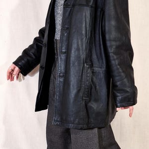 May include: A black leather jacket with a collared neckline and button closure. The jacket is worn over a gray turtleneck sweater and gray trousers. A pearl necklace is visible. The outfit is a vintage style.