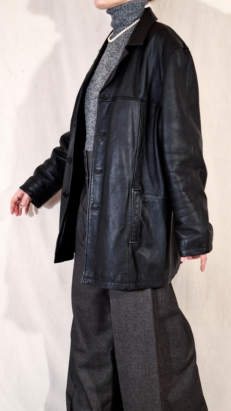 May include: A black leather jacket with a collared neckline and button closure. The jacket is worn over a gray turtleneck sweater and gray trousers. A pearl necklace is visible. The outfit is a vintage style.