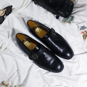 May include: A pair of black leather shoes with a buckle strap. The shoes are on a white surface with a black leather handbag, playing cards, and a pair of opera glasses.