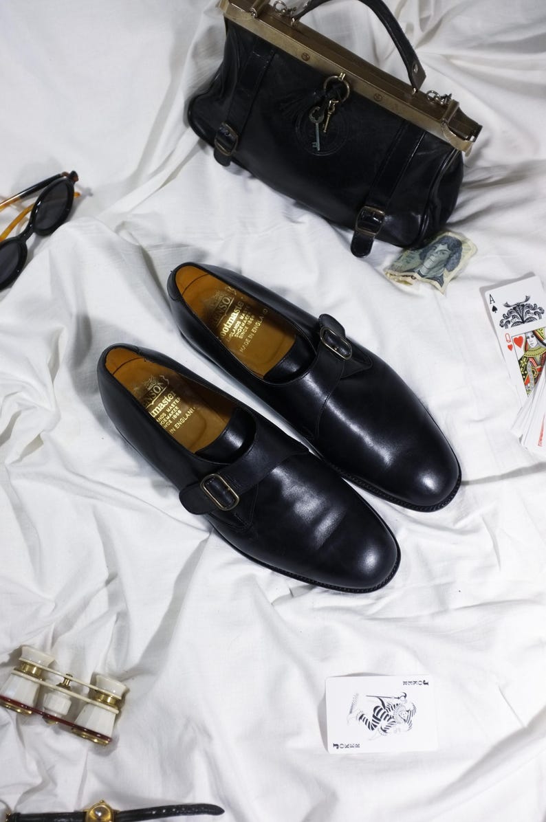 May include: A pair of black leather shoes with a buckle strap. The shoes are on a white surface with a black leather handbag, playing cards, and a pair of opera glasses.