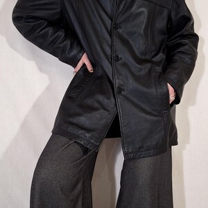 May include: A black leather jacket with a button-up front and a collared neckline. The jacket has pockets and is worn with gray pants. The outfit is completed with a gray turtleneck and a pearl necklace. The jacket appears to be a vintage style.