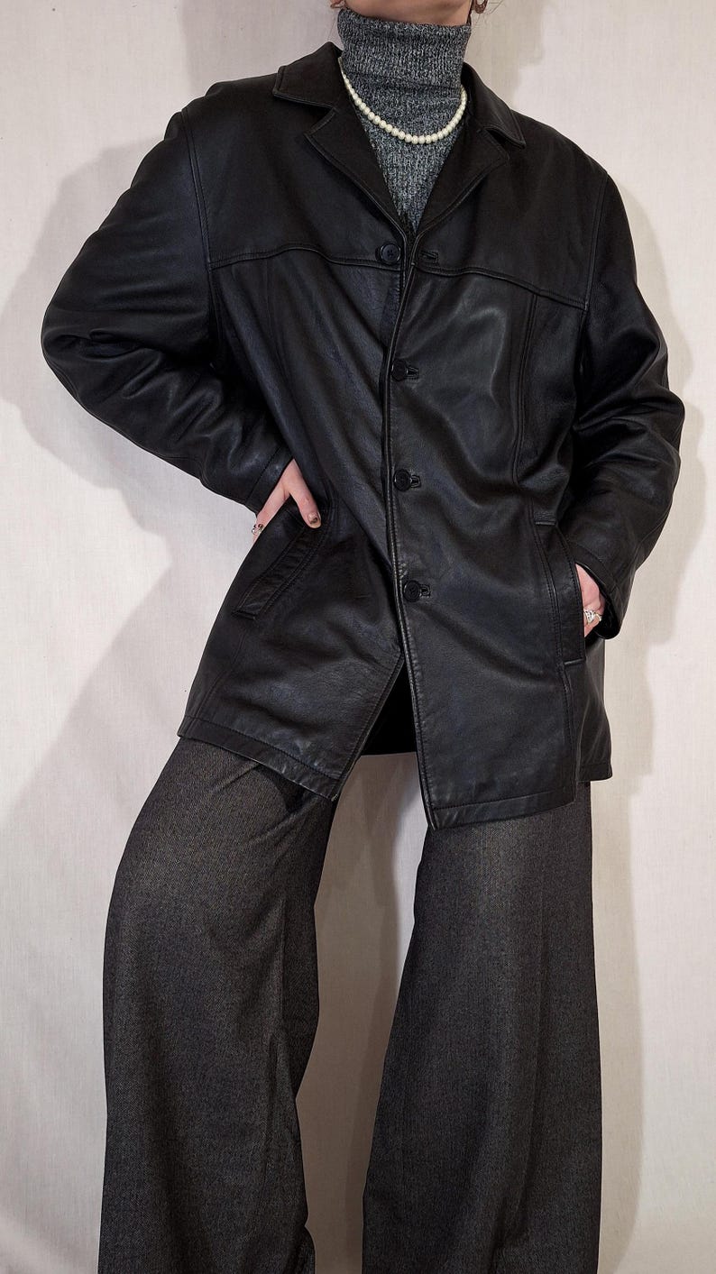 May include: A black leather jacket with a button-up front and a collared neckline. The jacket has pockets and is worn with gray pants. The outfit is completed with a gray turtleneck and a pearl necklace. The jacket appears to be a vintage style.