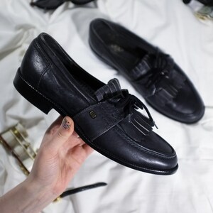 May include: A pair of black leather tassel loafers with a white stripe detail. The loafers have a rounded toe and a low heel.