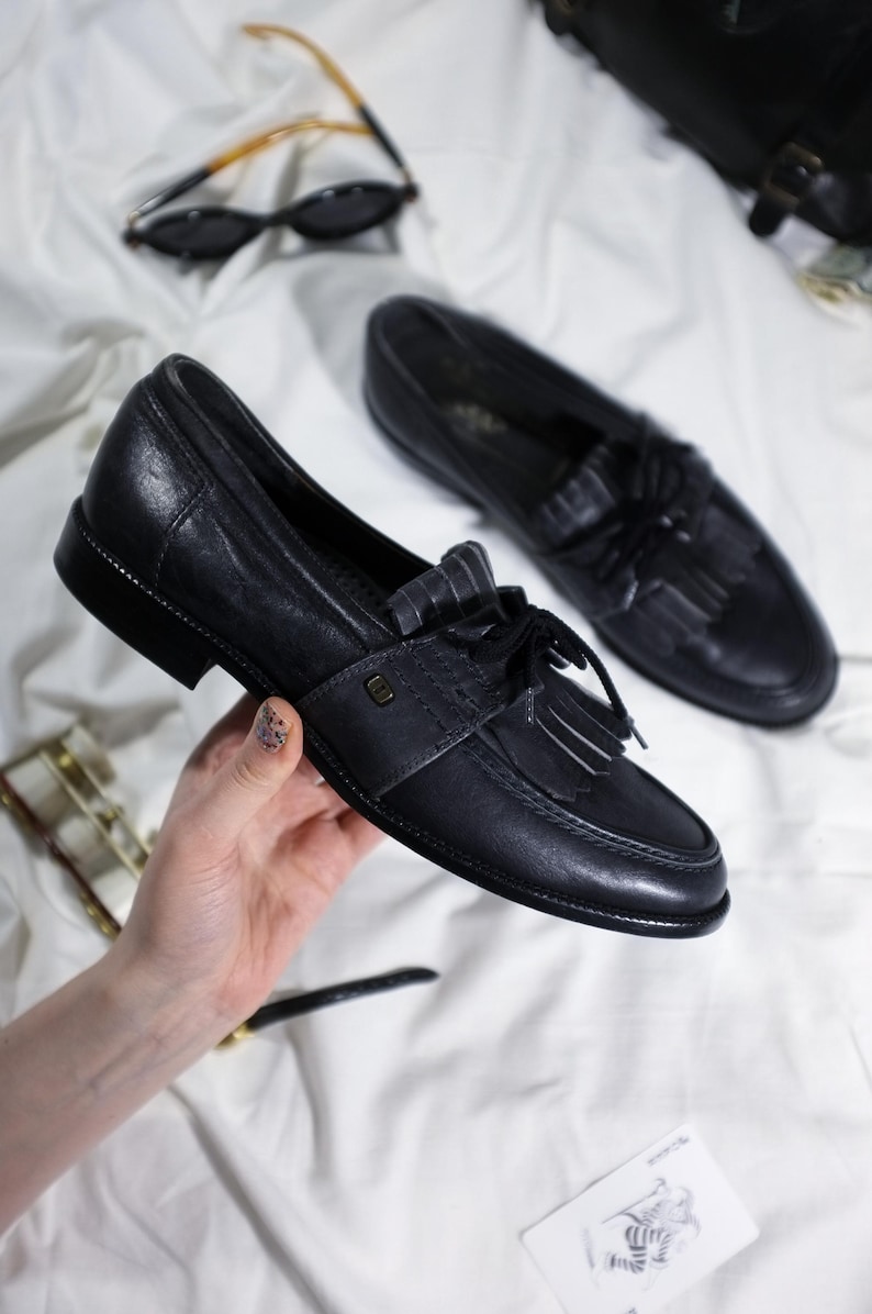 May include: A pair of black leather tassel loafers with a white stripe detail. The loafers have a rounded toe and a low heel.