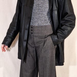 May include: A black leather jacket over a gray turtleneck sweater and high-waisted, wide-leg gray trousers. The jacket has a classic button-down design. A pearl necklace adds a touch of elegance to the outfit.