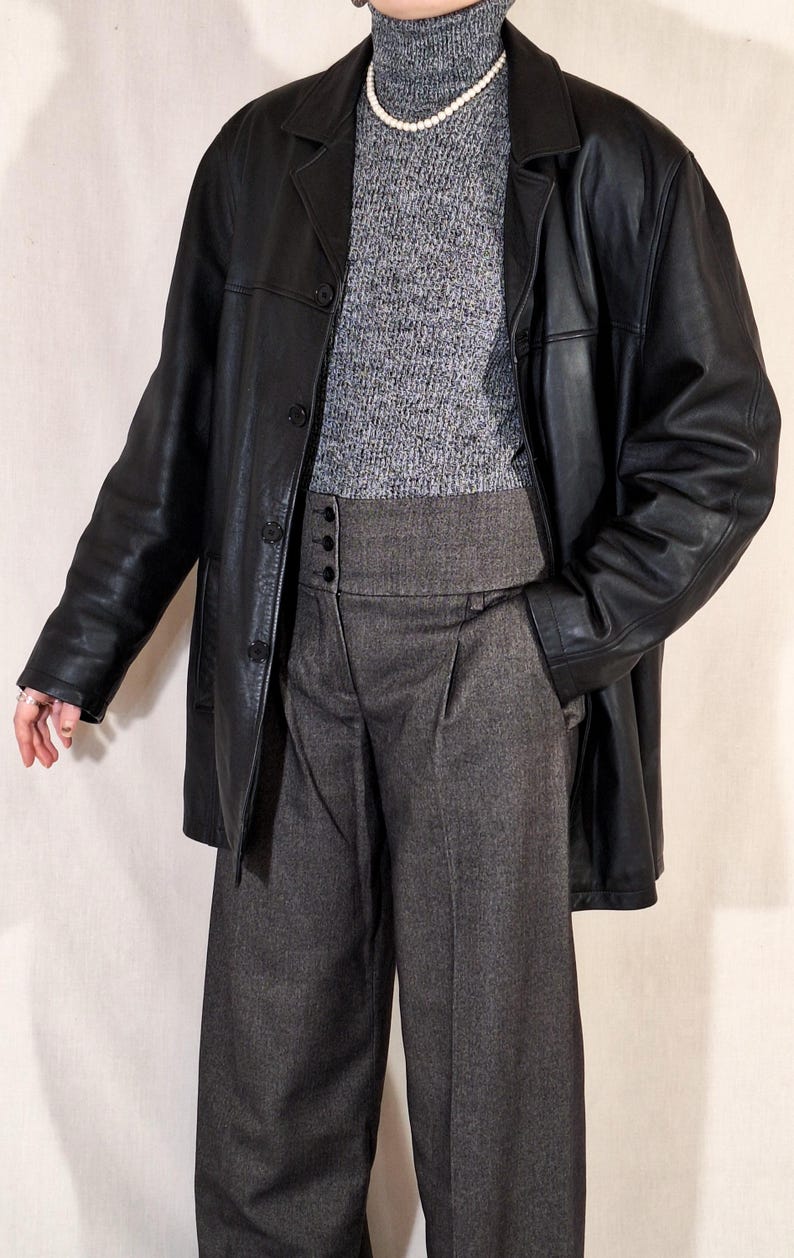 May include: A black leather jacket over a gray turtleneck sweater and high-waisted, wide-leg gray trousers. The jacket has a classic button-down design. A pearl necklace adds a touch of elegance to the outfit.