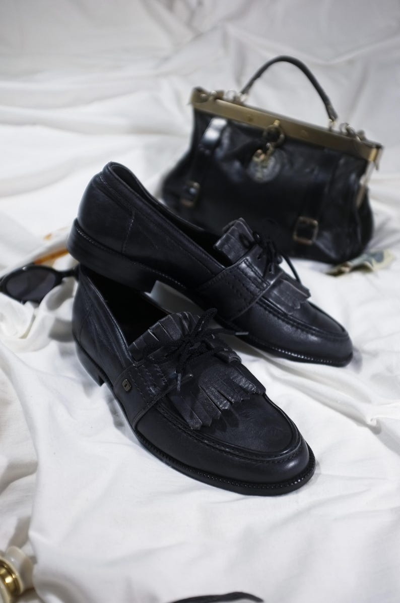 May include: A pair of black leather loafers with fringe detailing and a lace-up closure. The loafers are on a white surface.