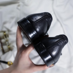May include: A pair of black leather shoes with a smooth finish. The shoes are held in a hand, showing the back of the shoes.
