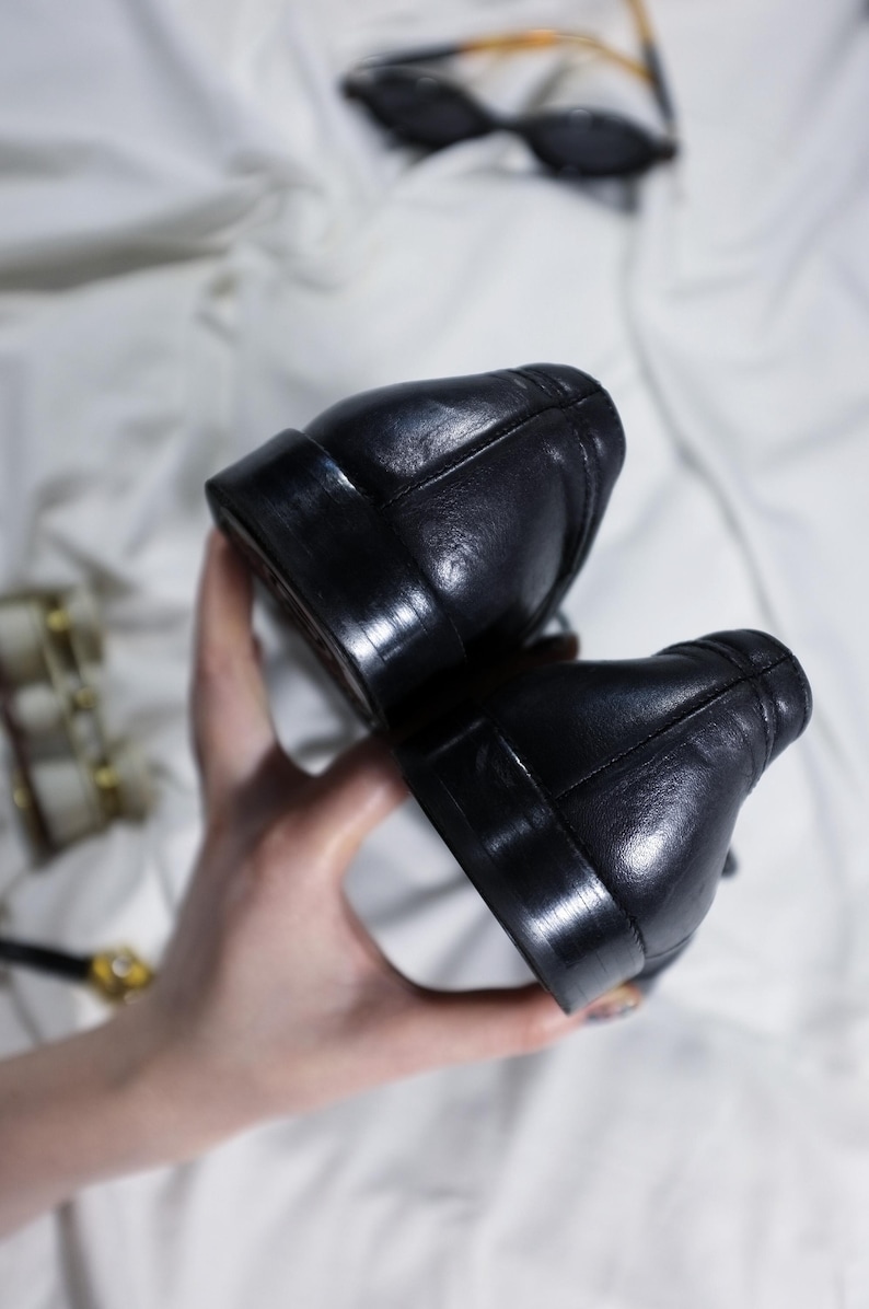 May include: A pair of black leather shoes with a smooth finish. The shoes are held in a hand, showing the back of the shoes.