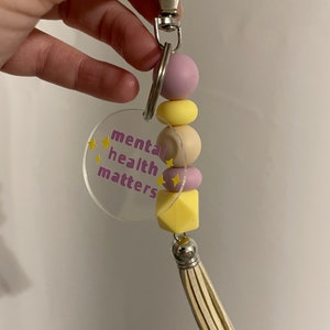May include: A keychain with a clear acrylic charm that says "mental health matters" in purple and yellow lettering. The charm is attached to a silver key ring with a purple, yellow, and beige beaded tassel.