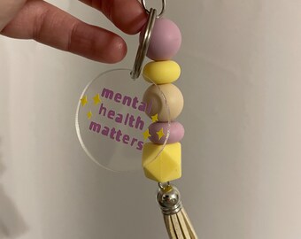 May include: A keychain with a clear acrylic charm that says "mental health matters" in purple and yellow lettering. The charm is attached to a silver key ring with a purple, yellow, and beige beaded tassel.