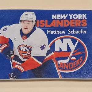 May include: A rectangular magnet featuring a hockey player in a white, blue, and orange New York Islanders jersey. The text reads NEW YORK ISLANDERS and Matthew Schaefer, accompanied by the team logo, a blue and orange circle with NY and a hockey stick.