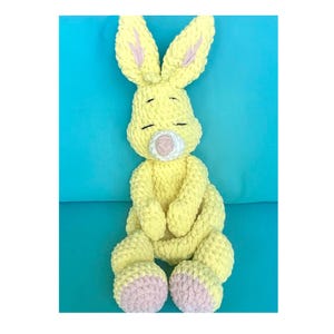 Puede incluir: Un conejito amarillo de ganchillo hecho a mano con orejas largas, ojos cerrados y nariz rosa. El interior de las orejas y las plantas de los pies del conejito son rosas. El peluche est&aacute; sentado sobre una superficie azul.