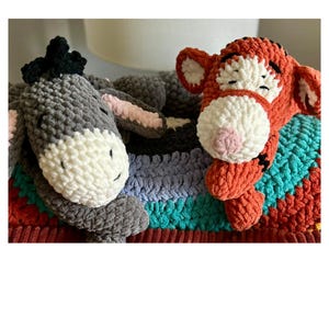 Puede incluir: Juguetes de peluche de ganchillo de un burro gris y un tigre naranja descansando sobre una manta a rayas. El burro tiene una cara blanca y orejas rosas, mientras que el tigre tiene un hocico blanco y rayas negras. La manta tiene rayas rojas, turquesas y azules.