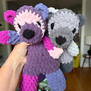 May include: Two handmade plush toys crafted from chunky yarn. One toy features a purple body with pink accents and a dark purple head. The second toy is primarily grey with a lighter grey head. Both have black stitched noses and eyes.