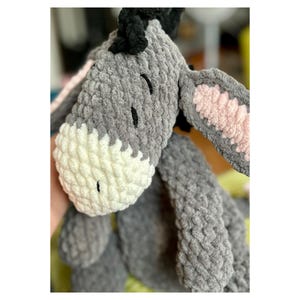 Puede incluir: Un juguete de peluche de burro gris y blanco hecho a mano con ojos negros y una melena negra. Las orejas del burro tienen detalles internos rosas. El juguete est&aacute; hecho de un material suave y texturizado.