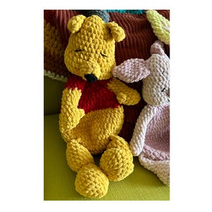 Puede incluir: Un peluche de Winnie the Pooh amarillo hecho a mano con una camisa roja y una nariz negra, junto a un peluche rosa. Los juguetes est&aacute;n hechos de un hilo suave y texturizado. Los peluches est&aacute;n sobre una superficie verde.