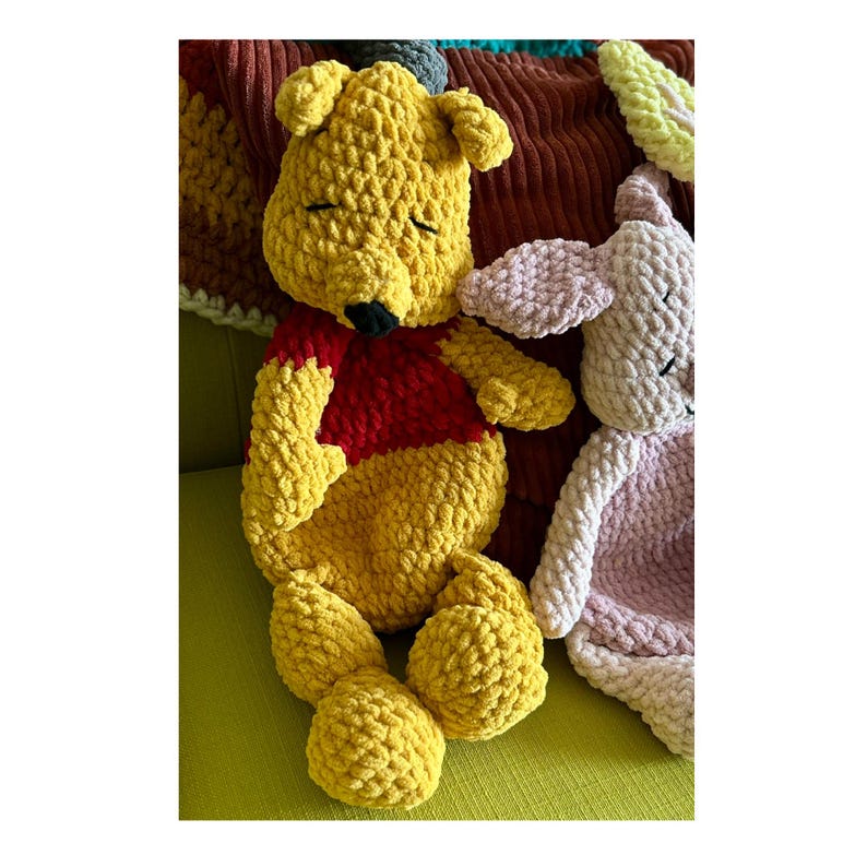 Puede incluir: Un peluche de Winnie the Pooh amarillo hecho a mano con una camisa roja y una nariz negra, junto a un peluche rosa. Los juguetes est&aacute;n hechos de un hilo suave y texturizado. Los peluches est&aacute;n sobre una superficie verde.