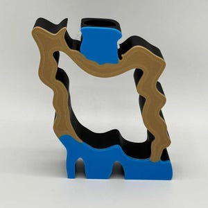 May include: A multi-layered, abstract sculpture in black, tan, and blue. The design features a wavy, organic shape with a rectangular blue element at the top and bottom. The sculpture is set against a plain white background.