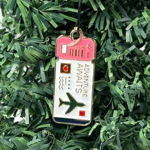 May include: A gold-colored metal ornament shaped like an airline ticket with the text "Adventure Awaits" and a small airplane graphic. The ticket is pink and white with a red heart.