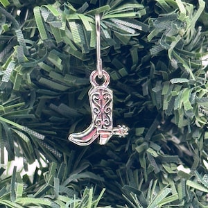 May include: Silver cowboy boot Christmas ornament with a spur. The boot has a detailed design and is hanging from a silver hook.