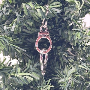 May include: Silver Christmas ornament shaped like handcuffs with the word "FREEDOM" inscribed on the top. The ornament is hanging on a green artificial Christmas tree.