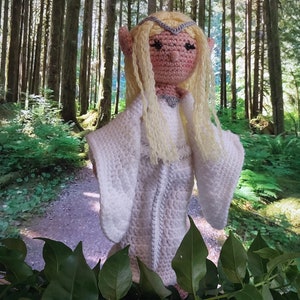 May include: A crocheted elf doll with long blonde hair, wearing a white gown and a silver crown. The doll is standing in a forest setting.