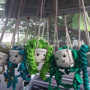 May include: Five crocheted hanging plant decorations with smiling faces. Each plant is a different color: green, teal, and white. The plants are hanging from a car's rearview mirror.