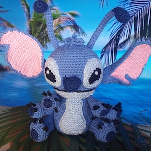 Könnte beinhalten: Ein blaues, gehäkeltes Stitch-Plüschtier mit rosa Ohren und schwarzen Augen. Das Spielzeug sitzt auf einer grünen, blättrigen Oberfläche.