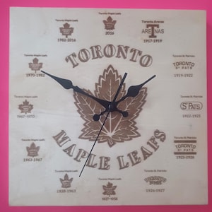 Puede incluir: Un reloj de madera con un diseño de hoja de arce y las palabras "Toronto Maple Leafs" alrededor del borde. La esfera del reloj muestra los años de la historia del equipo alrededor del borde.