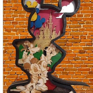 Puede incluir: Una obra de arte de madera en capas con la forma de la cabeza de Mickey Mouse. La obra presenta un castillo, globos y personajes de dibujos animados como Mickey, Minnie, Goofy y Donald Duck, sobre un fondo rosa y dorado. El marco es negro.