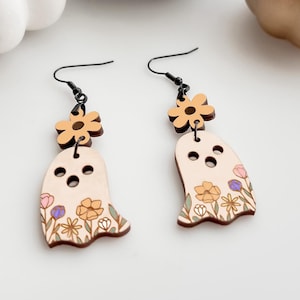 Könnte beinhalten: Ein Paar Ohrringe aus Holz in Form von Geistern mit Blumenmustern. Die Geister sind weiß mit gelben, rosa und violetten Blumen. Die Ohrringe sind an schwarzen Haken befestigt.